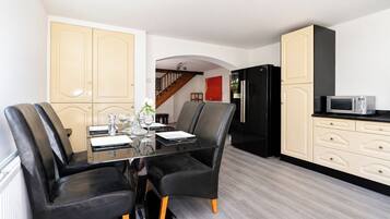 A dining area with a glass table and black chairs, a kitchen with a black refrigerator and white cabinets, and a wooden staircase in the background.
