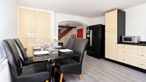 Un comedor con una mesa de vidrio y sillas negras, una cocina con un refrigerador negro y gabinetes blancos, y una escalera de madera al fondo.