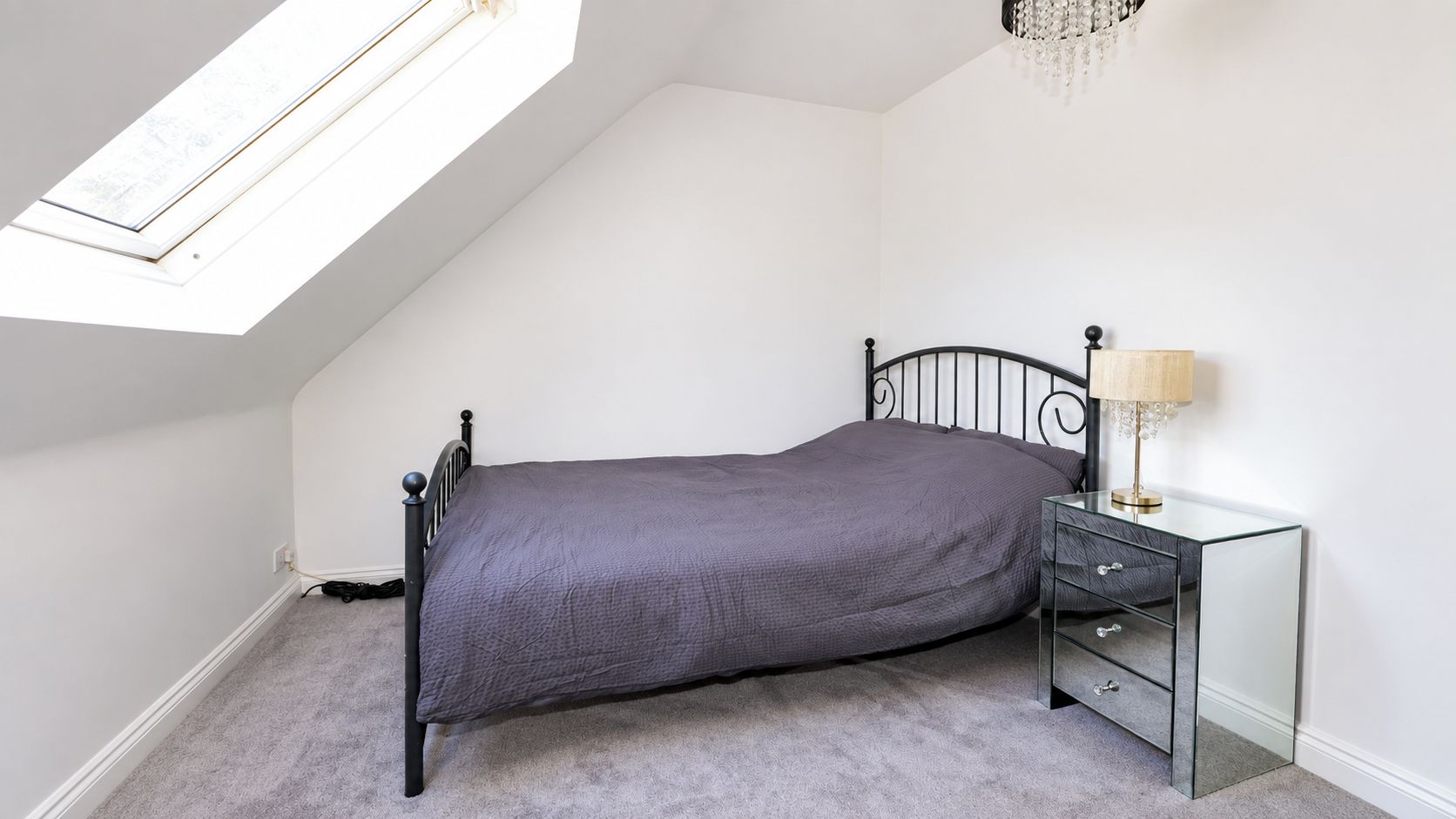 Une chambre avec un lit simple, une table de chevet avec des tiroirs, une lampe et une lucarne.