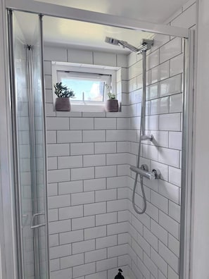 A shower with a glass door, white tiled walls, and a window with plants. - Cozy Cabin Retreat in Farnham (Farnham)