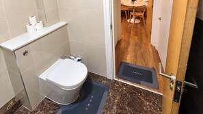 A bathroom with a toilet, a shelf with toilet paper, a shower area, and a wooden floor. - Acton Studio, Sleeps 2 with WiFi (London)