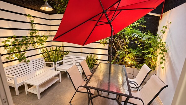 Un área de estar al aire libre con bancos y mesas blancos, un paraguas rojo y plantas en macetas.