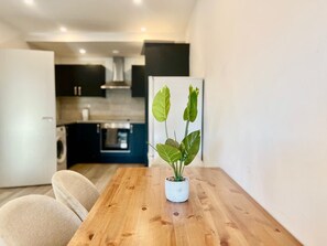 Uma mesa de jantar de madeira com uma planta em vaso ao centro, um sofá e uma cozinha ao fundo.