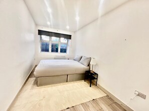 A bedroom with a bed, a nightstand, a lamp, a window, and a radiator. - Apartment Sleeps 5 near HIgh Street (Croydon)