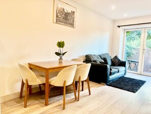 A modern living room with a wooden dining table, four chairs, a sofa, and a large window with a view of greenery. - Apartment Sleeps 5 near HIgh Street (Croydon)