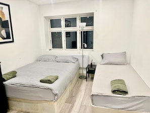 A room with two beds, a window, a floor lamp, and a framed picture on the wall. - Apartment Sleeps 5 near HIgh Street (Croydon)