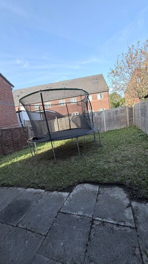 Un trampoline dans un jardin, avec une maison en briques en arrière-plan.