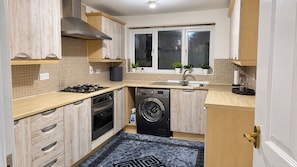 A compact kitchen with a gas stove, oven, washing machine, and a window above the sink. - Manchester Spacious 4-Bed House, Sleeps 6 (Manchester)