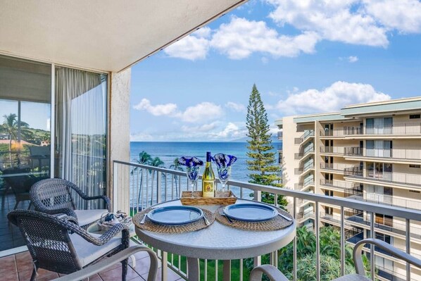 A balcony with a table set for two, overlooking a coastal view with a building in the background. - Updated oceanview 2 bedroom resort condo with AC, walk to shops, beach, & dining (Napili-Honokowai)