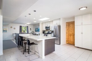 A modern kitchen with a central island, bar stools, a microwave, a refrigerator, and a wooden door. - Pickleball in Paradise | Sleeps 16, Free Pool Heat, Putting Green, Volleyball, & Games (Paradise Valley)