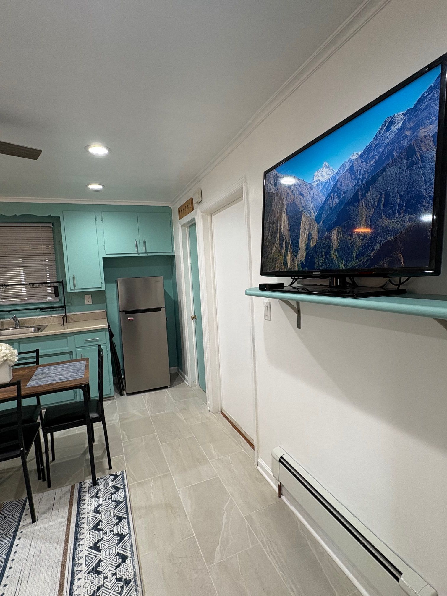 A modern kitchen with teal cabinets, a stainless steel refrigerator, a dining area with a table and chairs, and a mounted flat-screen TV displaying a mountain landscape.