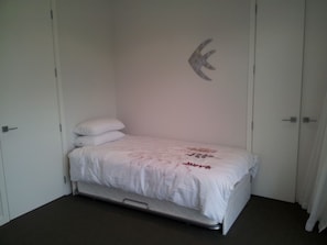Ein Einzelbett mit weißer Matratze und Kissen, ein kleiner Nachttisch und ein an der Wand befestigtes Regal mit einem dekorativen Gegenstand.
