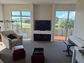 Ein Wohnzimmer mit Flachbildfernseher, einem grauen Sessel, einem roten Hocker, einer Bank und einem Schreibtisch mit Computer.