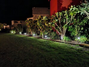 Ein Garten bei Nacht, gestaltet mit einer Wegeführung, gesäumt von kleinen Bäumen und Sträuchern, die durch Bodenleuchten erhellt werden.