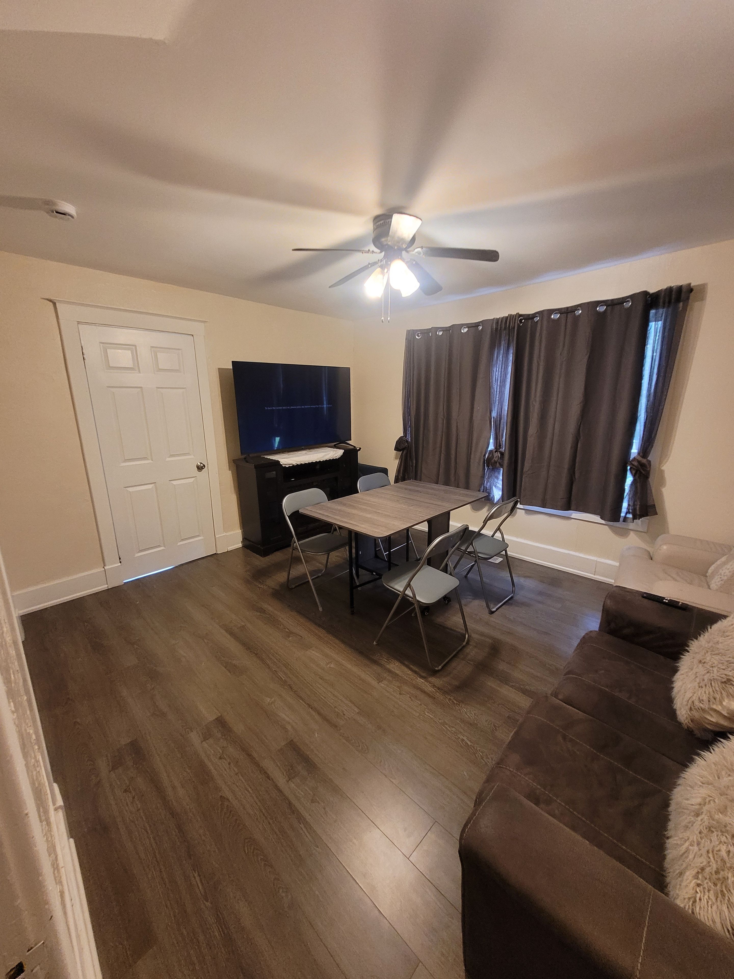 A living room with a ceiling fan, a flat-screen TV, a dining table with chairs, a sofa, and a window with curtains.