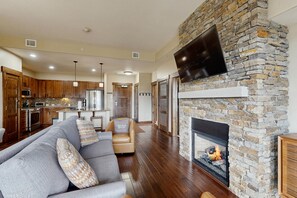 A spacious living room with a stone fireplace, a flat-screen TV mounted on the wall, and a kitchen in the background. - Trailhead Lodge #5116 condo with fireplace, under three miles from downtown (Steamboat Springs)