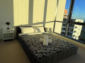 A bedroom with a large window, a bed with a patterned bedspread, a nightstand with a lamp, and a view of buildings outside. - Panoramic Ocean 2BR, Gold Coast Spa & Pool (Southport)
