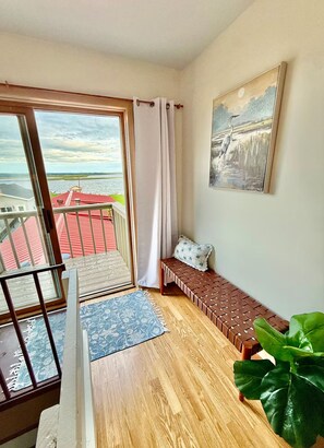 Quarto com varanda, piso de madeira, um banco, uma planta grande e um quadro na parede.