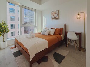 A modern bedroom with a large bed, a bedside table, a chair, a potted plant, and a window with a view of buildings. - Cozy 2BR • Gym+Parking • Rogers Centre | MTCC (Toronto)