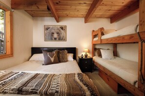 Ein Zimmer mit einem großen Bett, einem Holzbett, einem Nachttisch mit Lampe und einem Bärenbild an der Wand.
