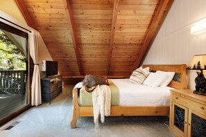 A bedroom with a sloped ceiling, a bed with pillows and a blanket, a bedside table with a lamp, and a view of the outdoors through a sliding door. - Cozy 3-bedroom cabin in superb Lake Arrowhead (Lake Arrowhead)