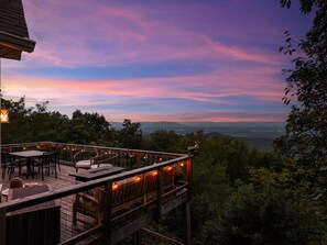 A wooden deck with outdoor furniture and string lights, overlooking a forested landscape and distant city lights. - Summit Shenandoah | Valley Views, Hot Tub, Sauna + Theater | 4BR, Sleeps 8 (Skyland Estates)