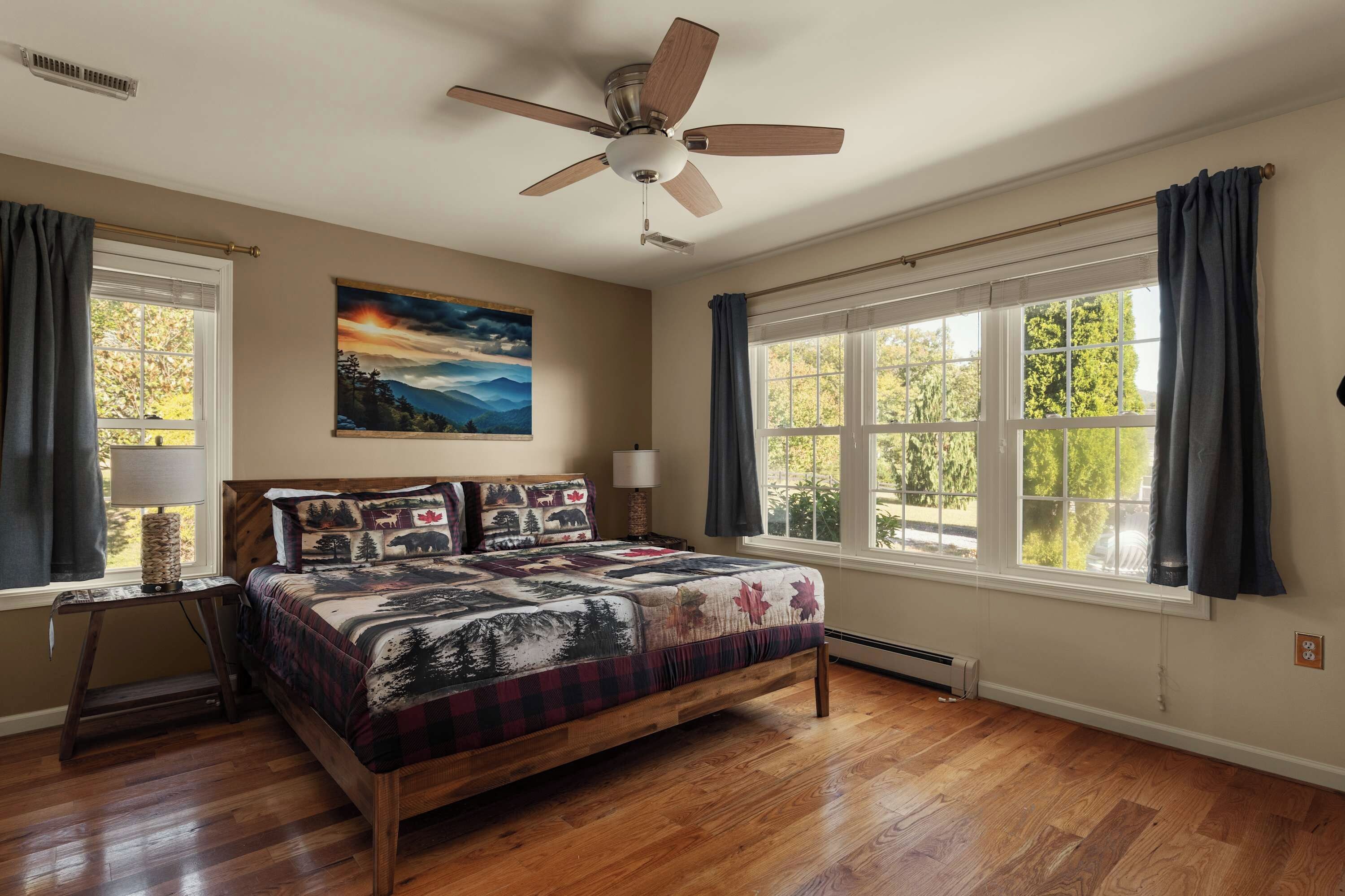 A bedroom with a bed, a ceiling fan, a painting, and large windows.