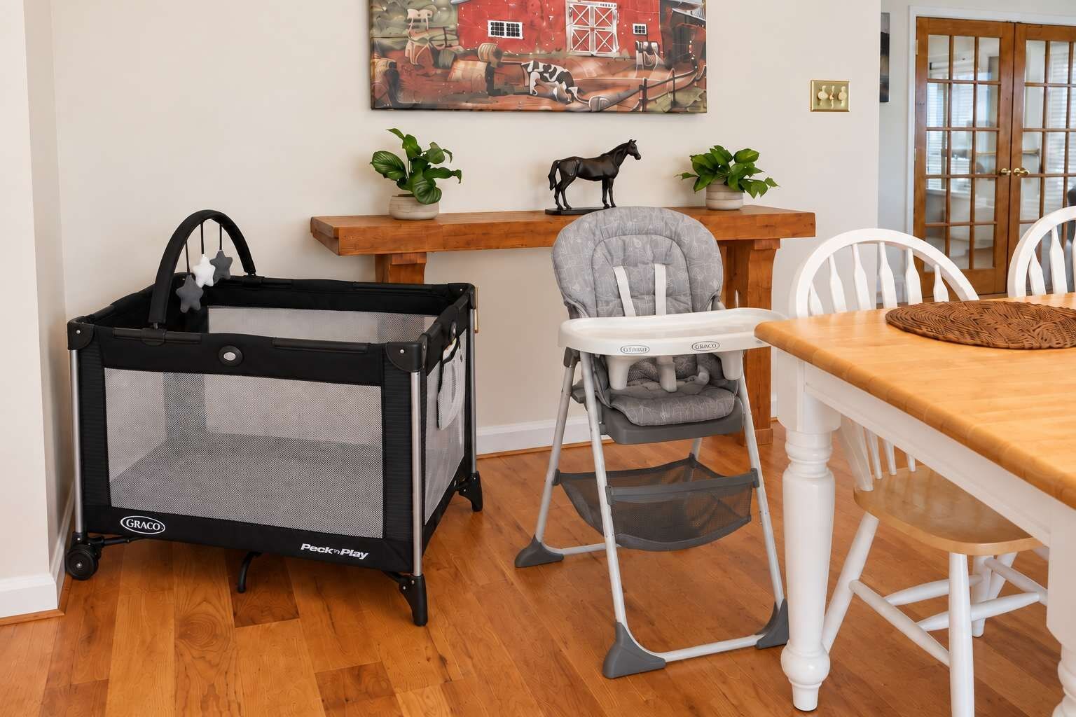 Een kamer met een houten eettafel, een kinderstoel, een babybedje en een schilderij aan de muur.