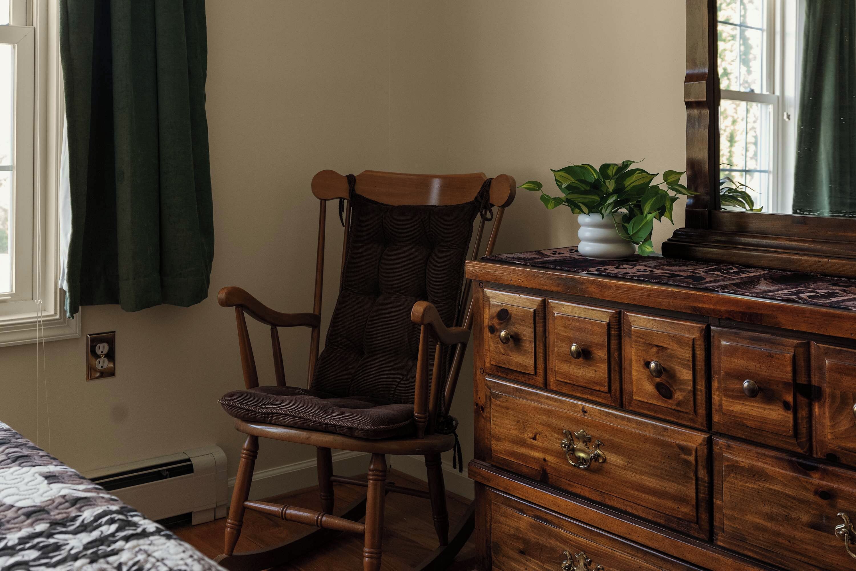 Una habitación con una mecedora de madera, un aparador con varios cajones, una planta en maceta y un ventanal con cortinas.