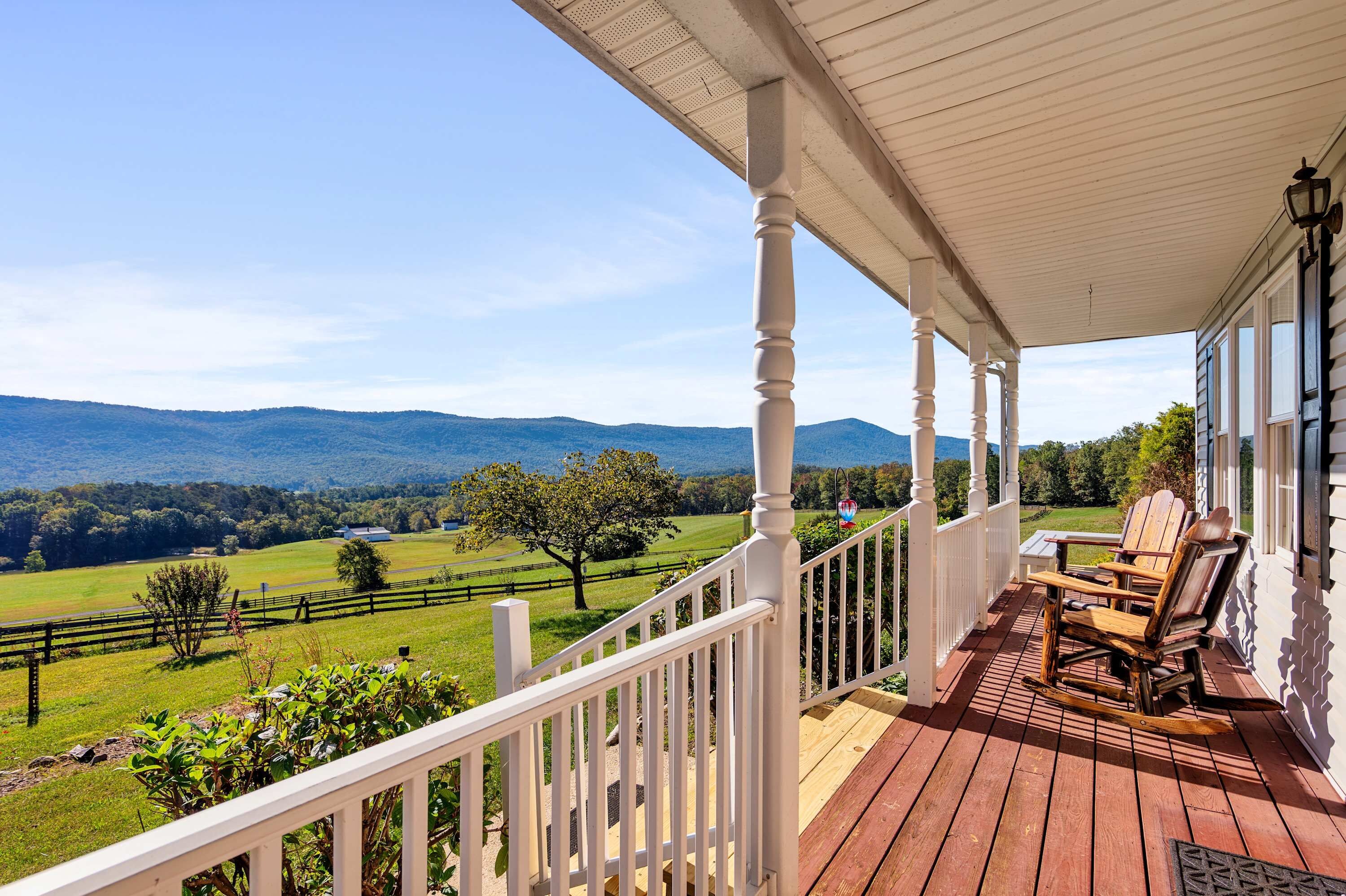 A wooden deck with outdoor furniture, a white railing, and a view of a green landscape with mountains in the distance.