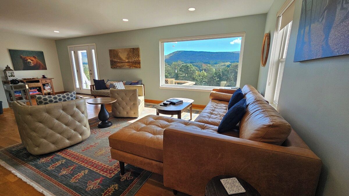 A living room with a sofa, armchairs, a coffee table, and a rug. There is a window with a view of a mountain landscape.