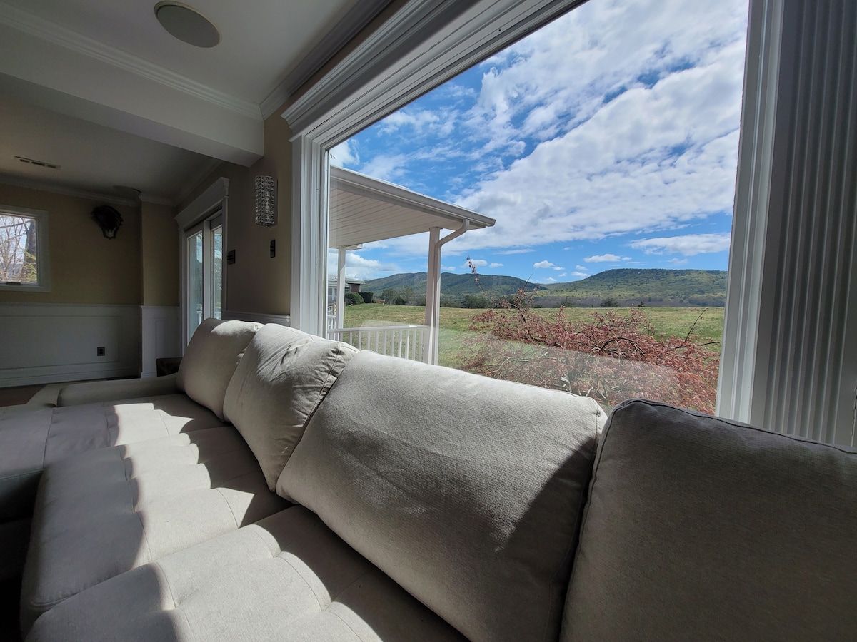 A beige sofa in a room with a large window overlooking a landscape with hills and a clear sky.