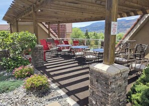 An outdoor patio with tables and chairs, a stone pillar, and a view of mountains. - Downtown Waterfront  | 2BR/2BA, Pool & Free Parking (Kelowna)