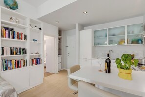 A modern kitchen with white cabinetry, a dining area with a beige chair, and a bookshelf filled with books. - Park Apt With AC & Private Balcony, 300 mt to Sea, Torre del Lago, Italy (Torre del Lago)
