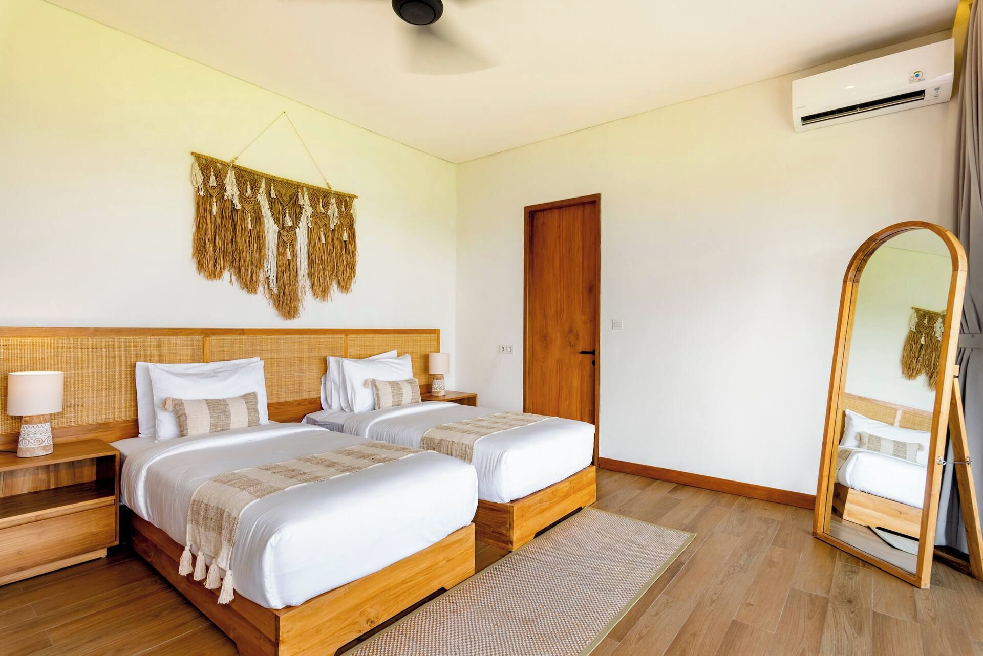 Habitación de hotel con dos camas, cabecero de madera, aire acondicionado de pared, tapiz de pared tejido, puerta de madera y espejo de cuerpo entero.