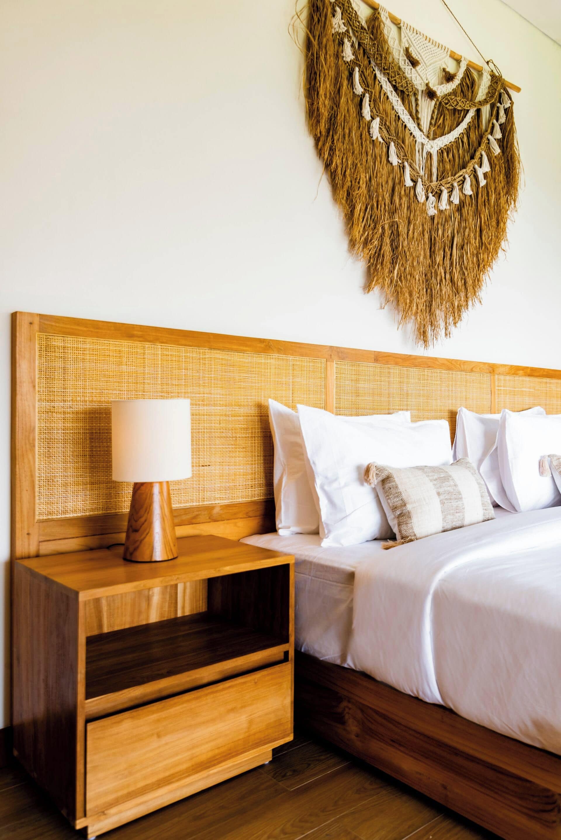 Un dormitorio con cabecera de madera, mesita de noche con lámpara y un colgador de macramé en la pared.
