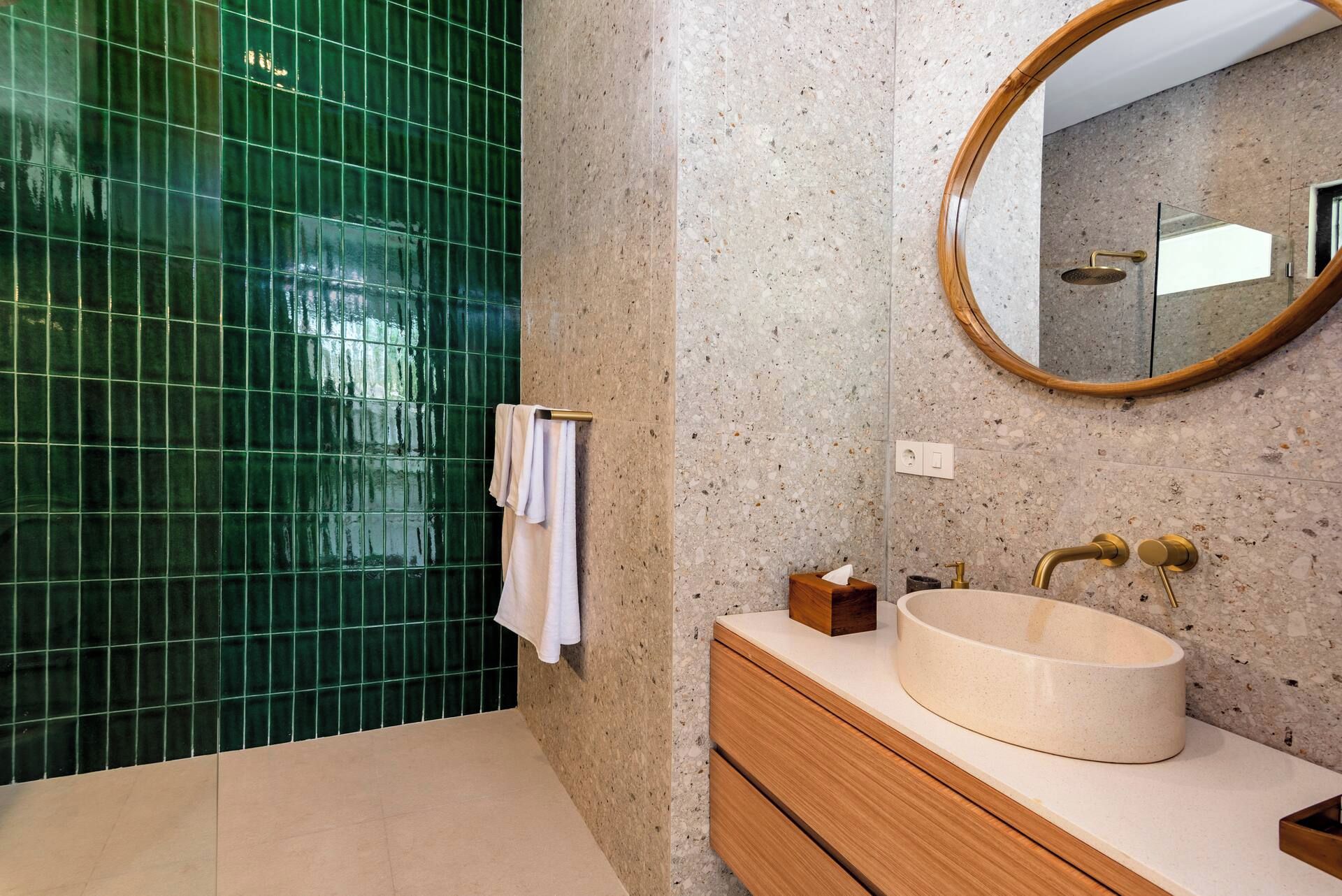 A bathroom with a green tiled wall, a round mirror, a beige sink, and wooden cabinetry.