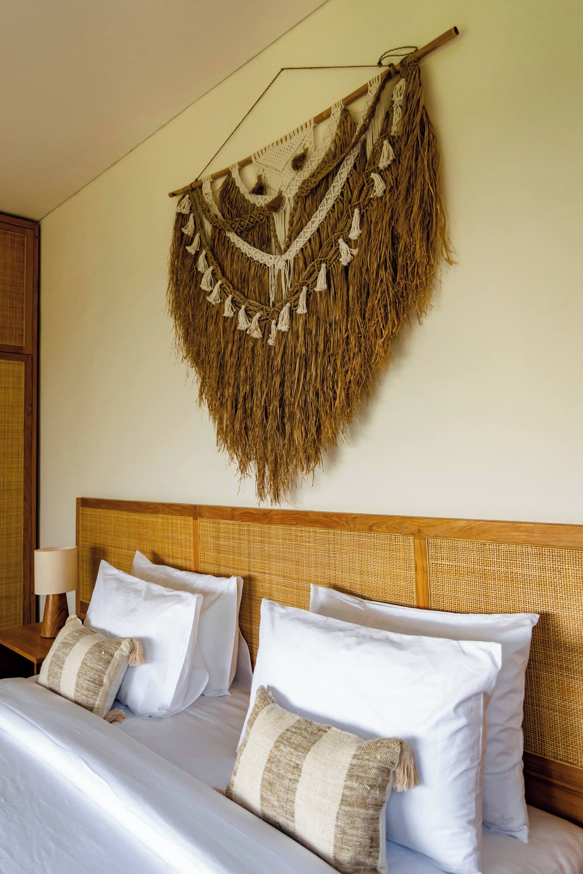 Una cama con sábanas blancas y dos almohadas, cabecera de tejido y un tapiz con flecos.