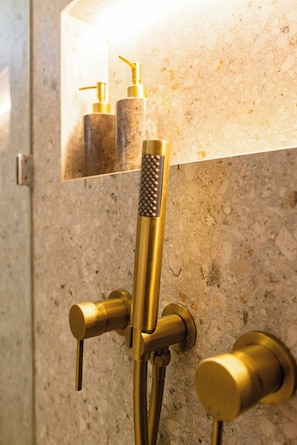 Un baño con encimera de granito, ducha con cabezal dorado y un espejo que refleja un dispensador de jabón.