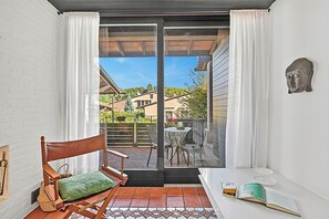 A small room with a sliding glass door leading to a patio. Inside, there's a wooden chair with a cushion, a small table with a book and a vase, and a wall-mounted sculpture. - Borgo Della Motta Swimming Pool &Golf, Travedona Monate, Italy (Travedona Monate)
