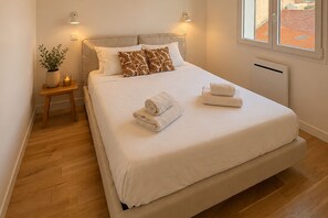 A neatly made bed with white linens and two rolled towels, a bedside table with a plant and candle, and a window with a view of buildings. - Sanary Apt N2, Sanary-sur-Mer, France (Sanary-sur-Mer)