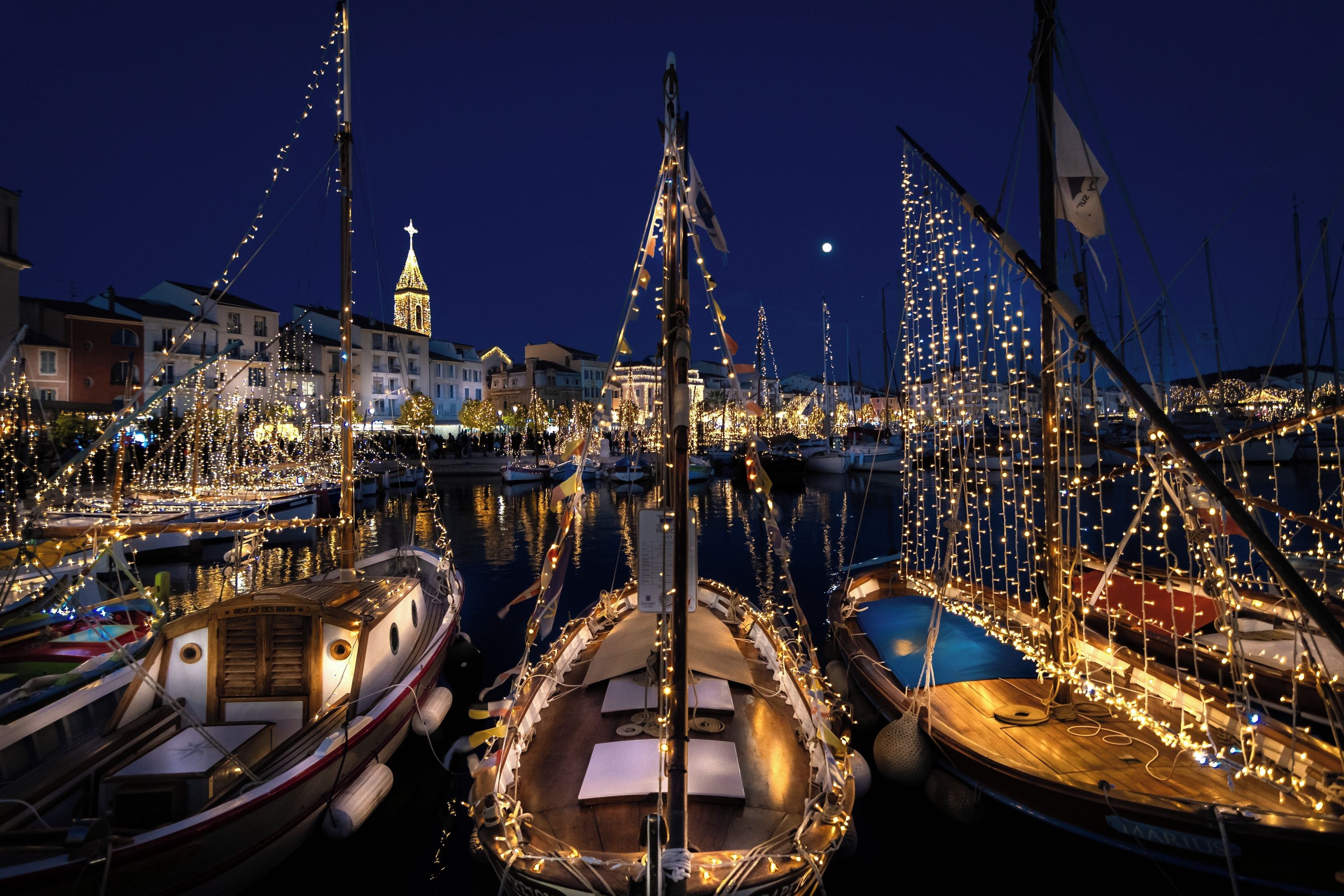 Un port de plaisance la nuit, avec des bateaux amarrés et décorés de lumières, une tour imposante en arrière-plan et un ciel nocturne dégagé.
