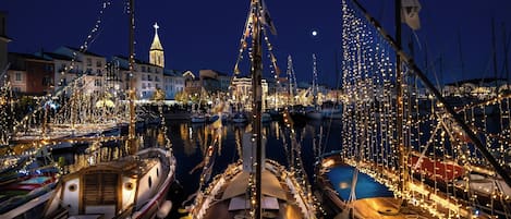A marina at night with boats docked and decorated with lights, a prominent tower in the background, and a clear night sky.