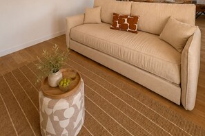 Un sofá color beige con dos almohadones decorativos, una mesita redonda de madera con un jarrón y un cuenco de aceitunas, y un tapete tejido.