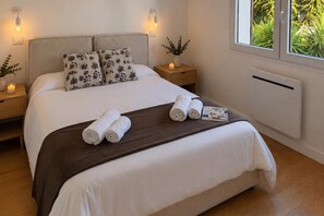 Un lit bien fait, avec du linge de lit blanc et un plaid marron, deux tables de chevet avec des bougies, une petite table de nuit avec un vase et une bougie, et une fenêtre donnant sur un paysage verdoyant.