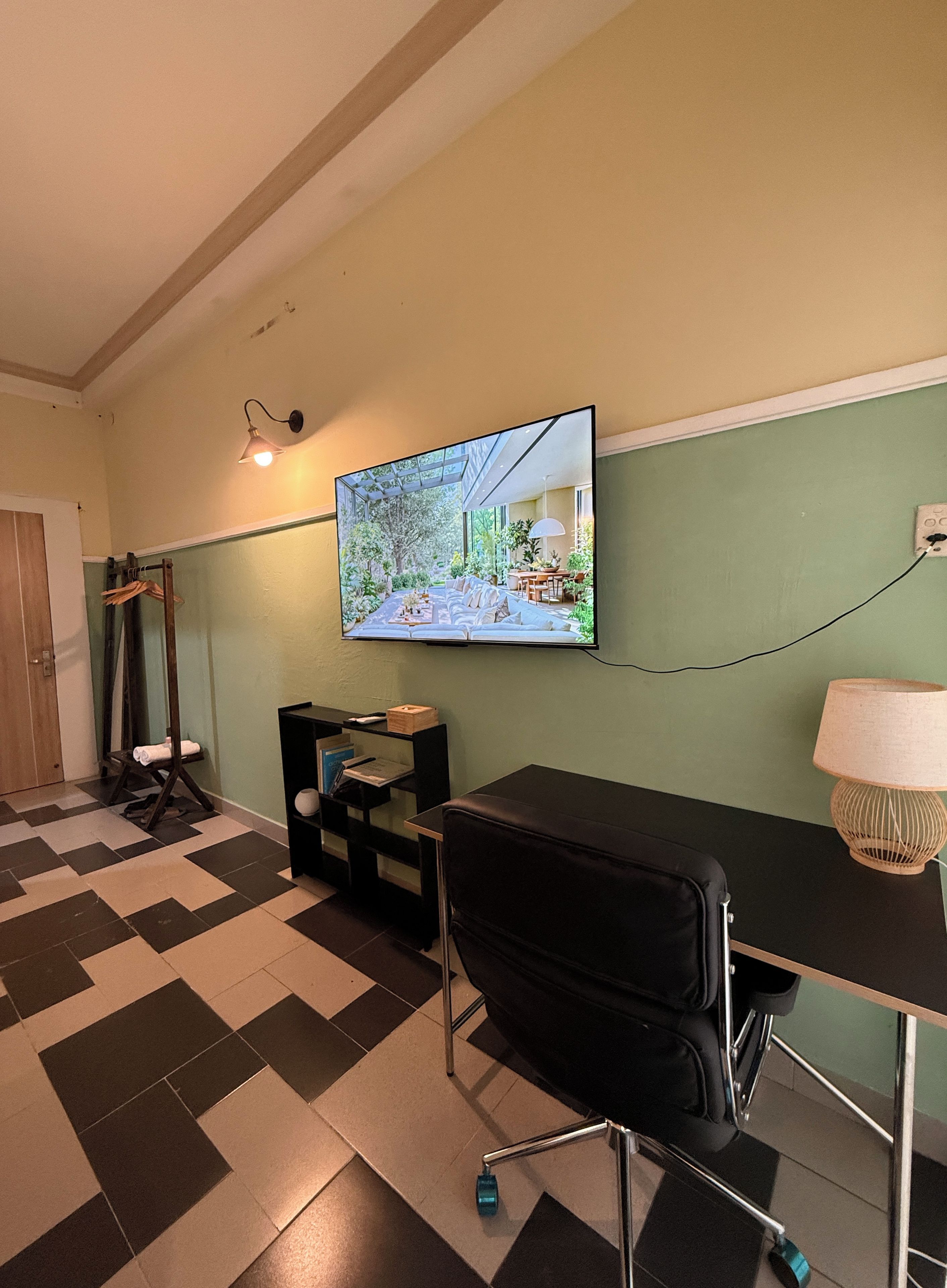 A room with a television mounted on the wall, a black office chair, a desk with a lamp, and a checkered floor.