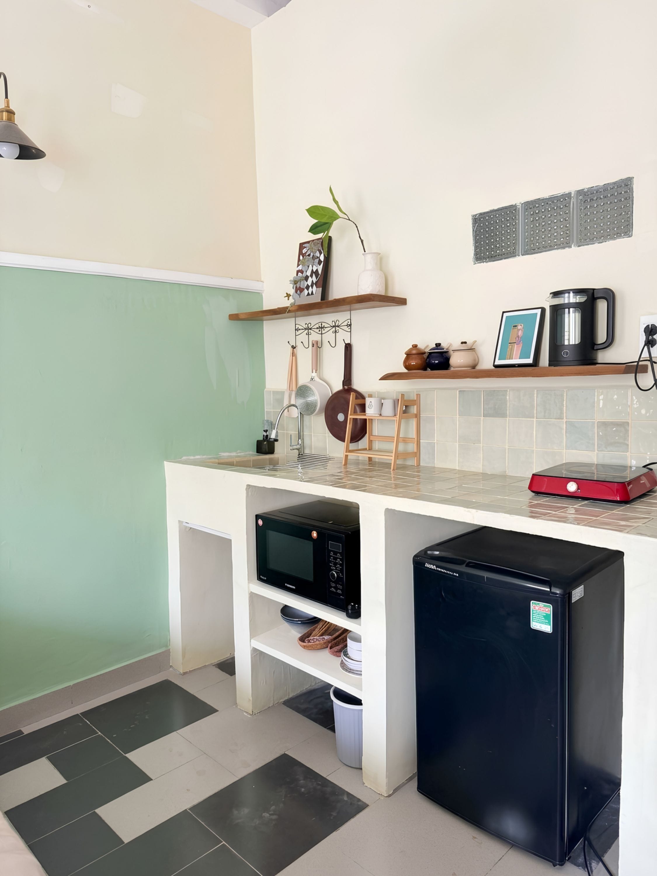 A compact kitchen with a microwave, refrigerator, and a small sink. There is a shelf with kitchenware and a hanging plant.