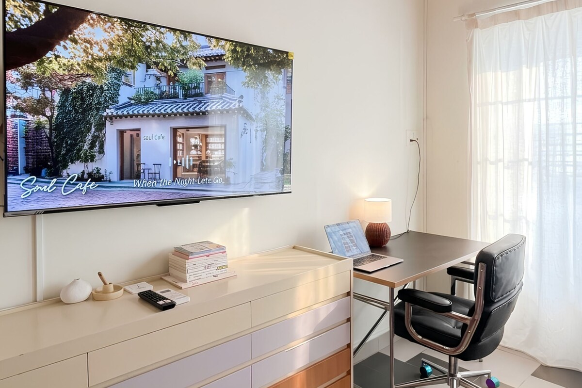 Una habitación moderna con una televisión de pantalla plana en la pared, un escritorio con una computadora portátil, una silla y una mesita con una lámpara.