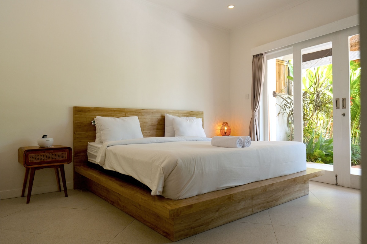 A bedroom with a wooden bed frame, white bedding, a bedside table with a lamp, and a view of greenery through a sliding door.