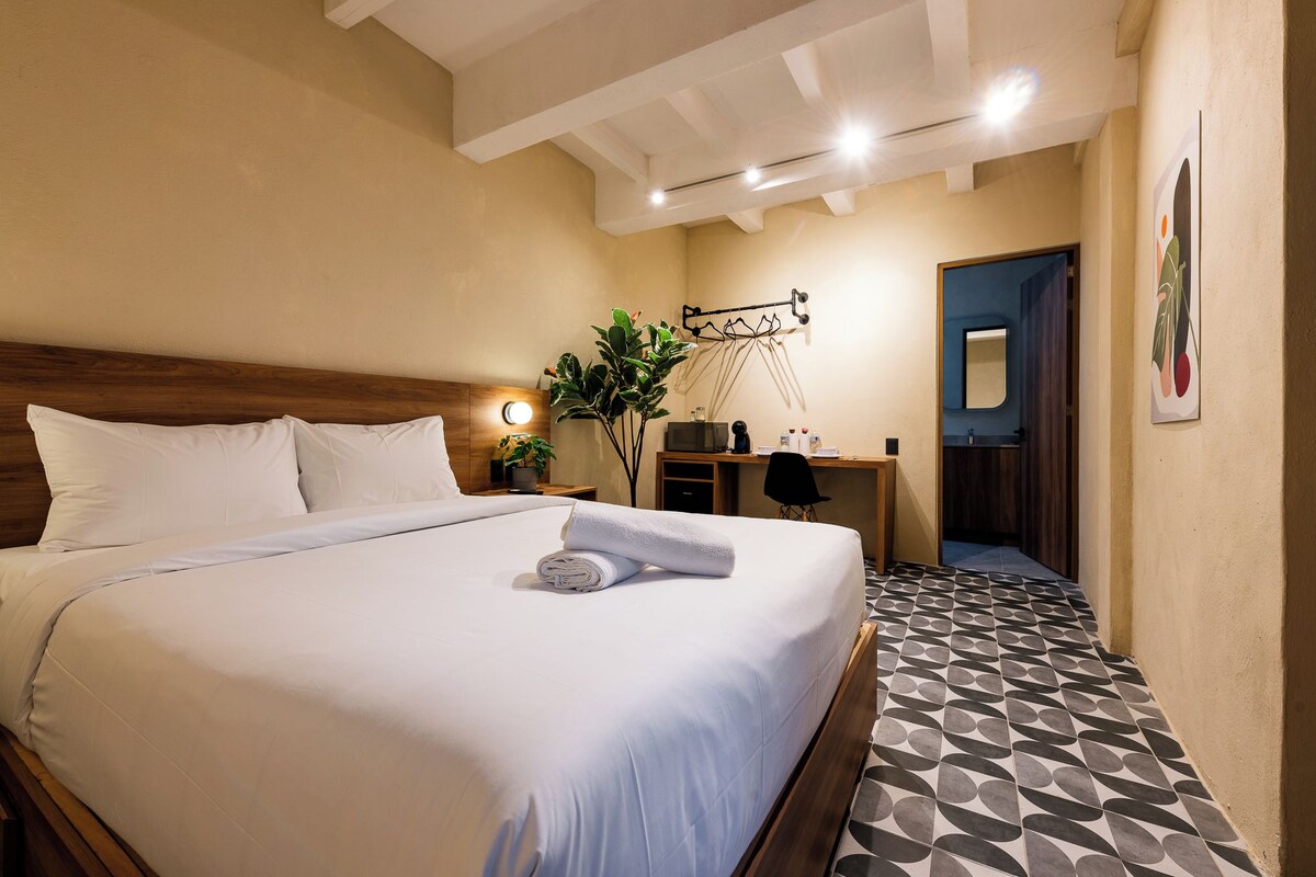 Una habitación de hotel con cama, mesita de noche, lámpara y una planta en maceta.